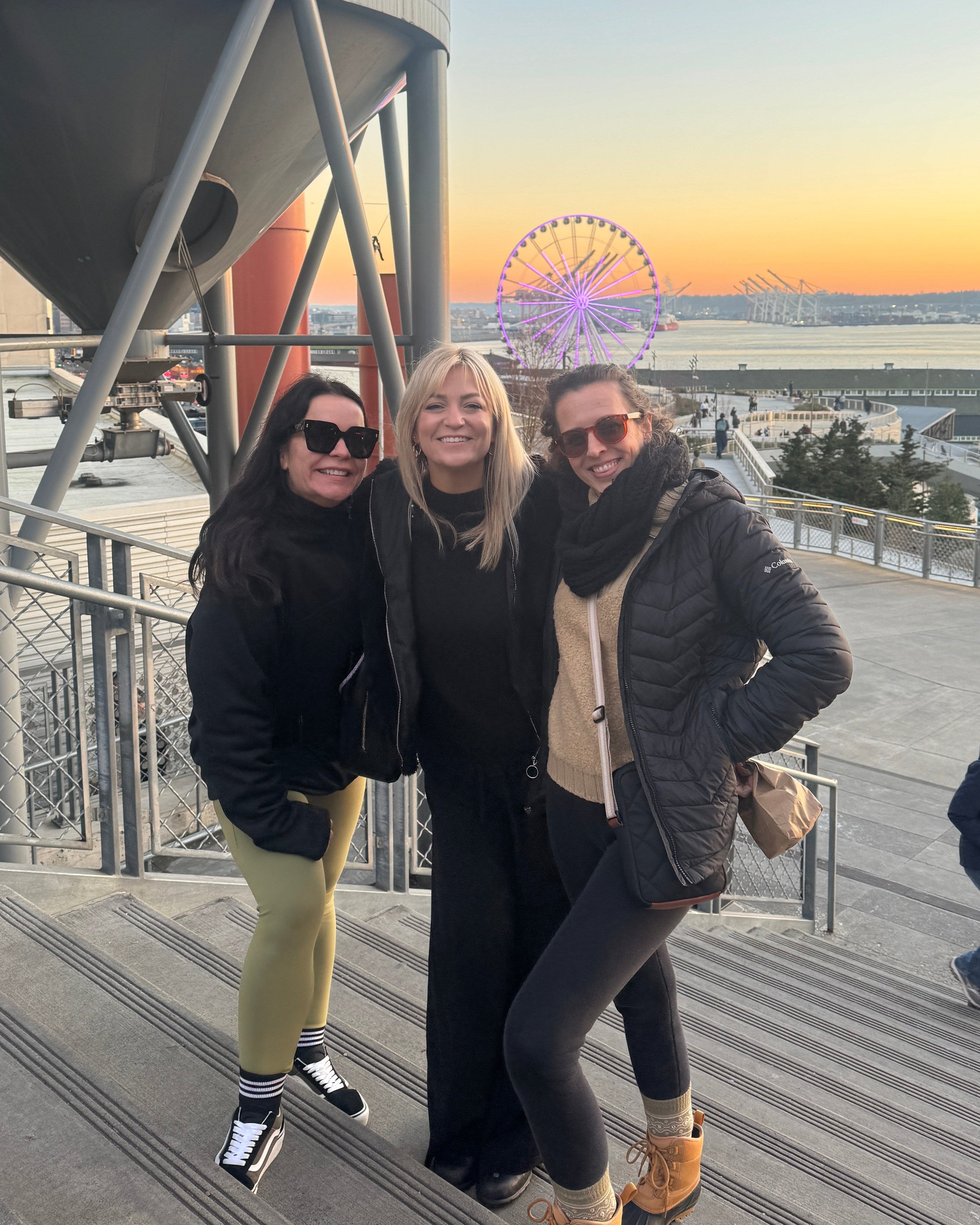 Three people posing for a photo with a Ferris wheel and sunset in the background.
