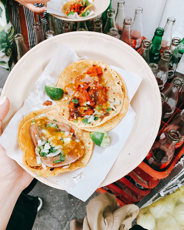 Two tacos on a plate held by a person with a background of bottles and food.