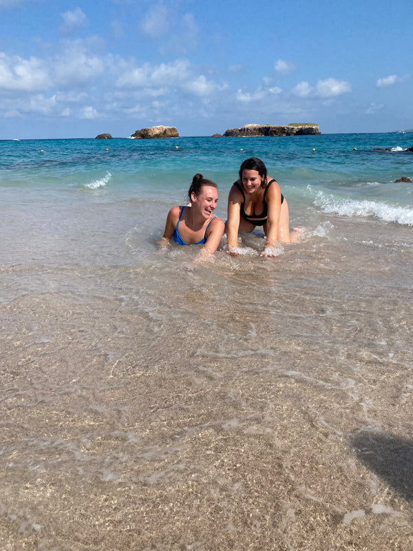 Two people sitting in shallow water at the beach with clear blue sky.