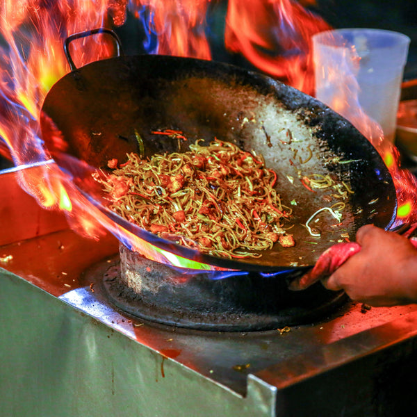 Person cooking food in a wok over an open flame on a stove.