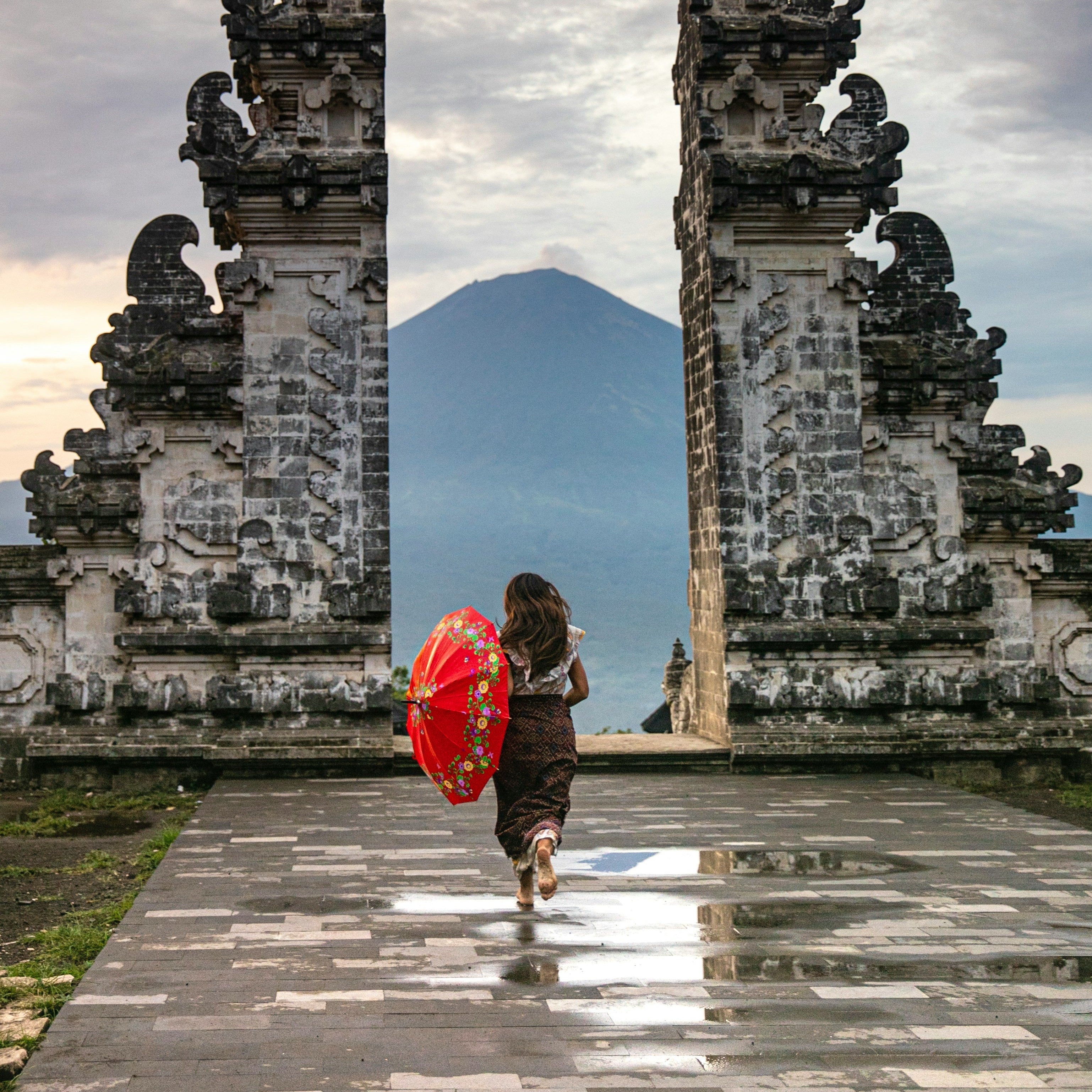 Person holding a red umbrella walking through stone temple ruins with a mountain in the background