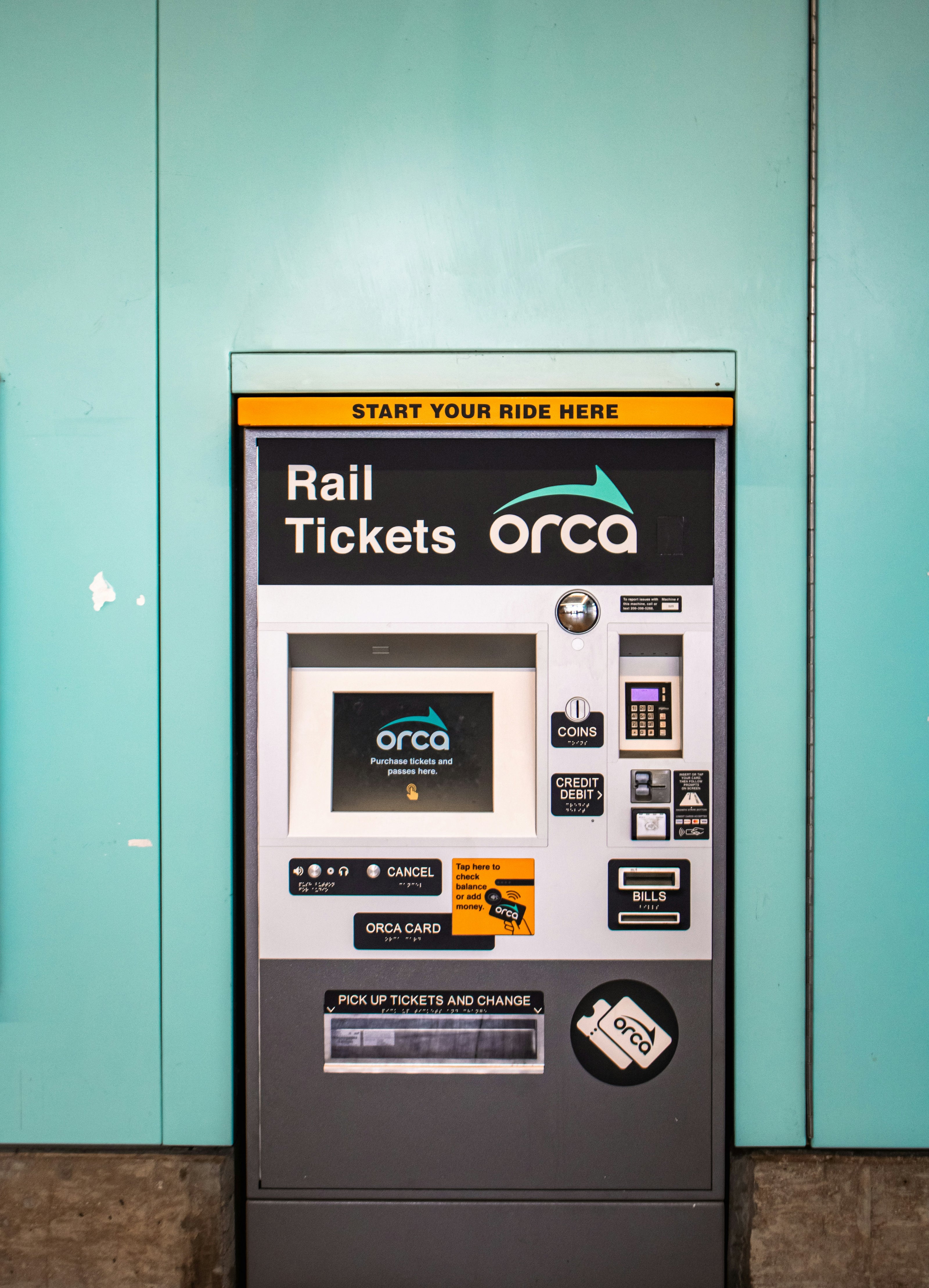 Vending machine for rail tickets with 'Orca' branding against a teal wall.