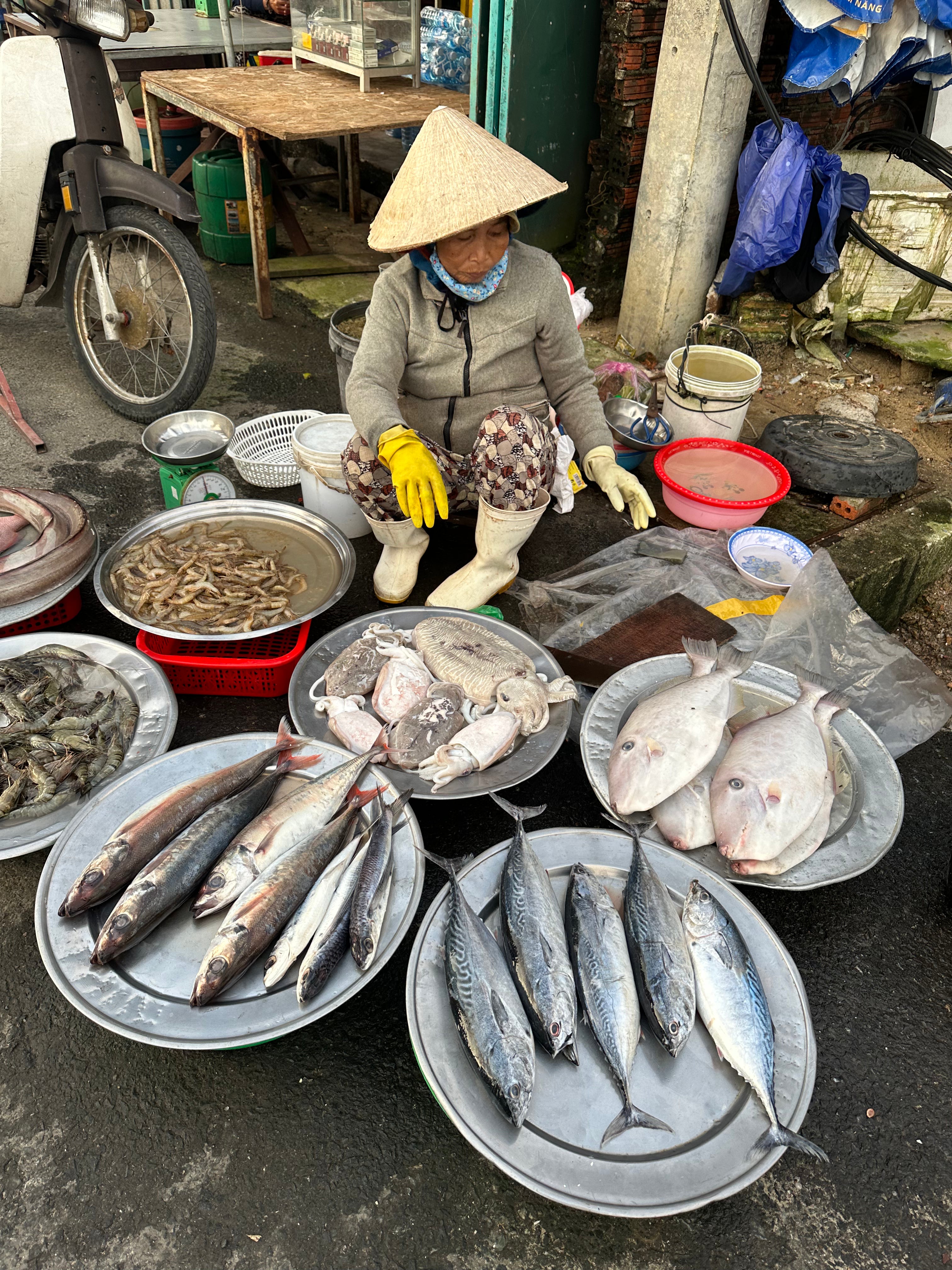 Woman selling fish at a market with various fish on trays and a motorcycle in the background.