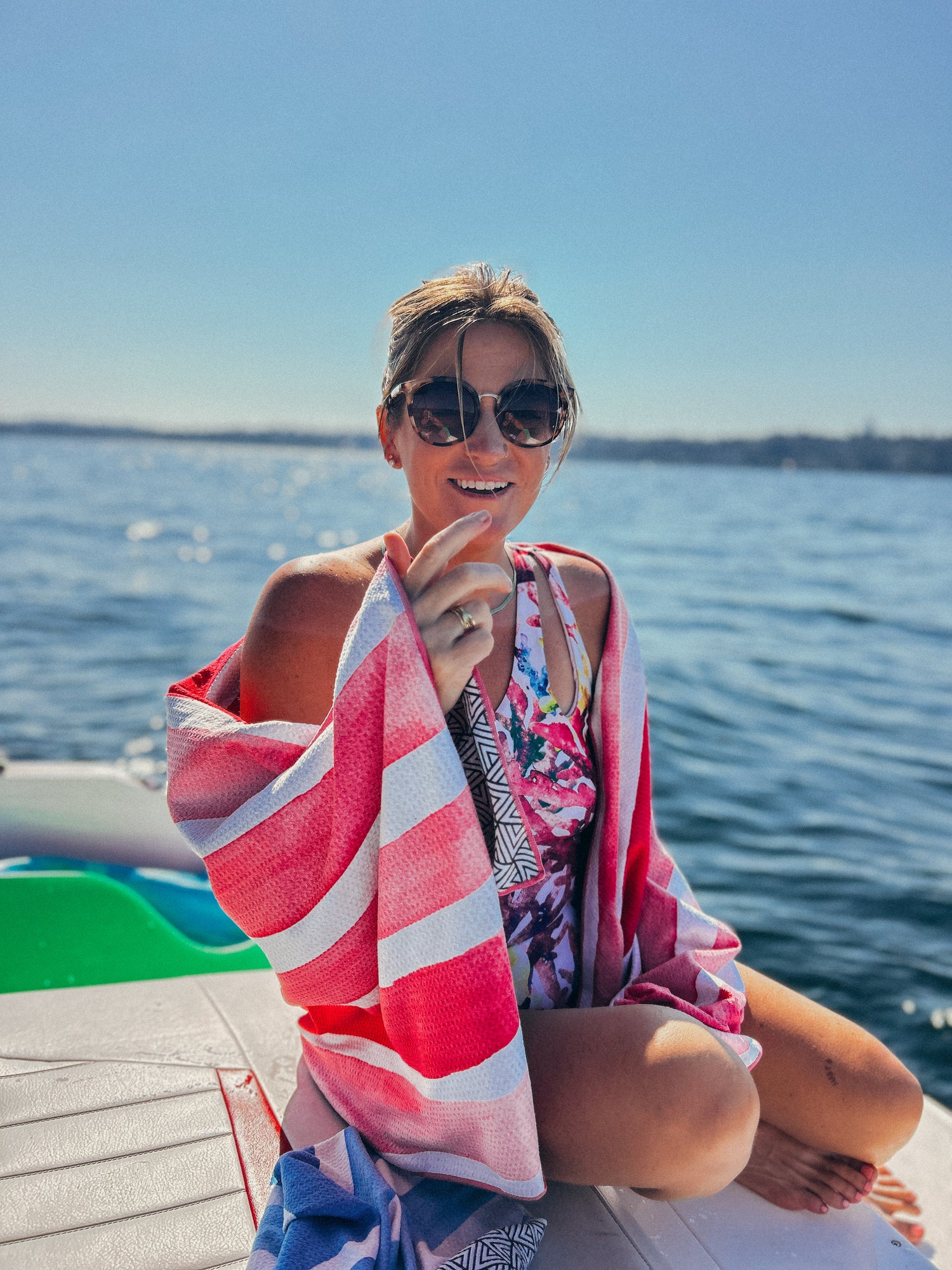 Woman sitting on a boat with a pink and white striped towel, wearing sunglasses and a colorful swimsuit.